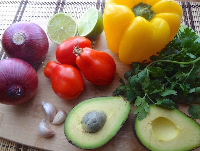 Potato Tacos ingredients photo. 2 whole red onions, a lime sliced in half, 3 garlic cloves, 3 roma tomatoes, a yellow bell pepper, an avocado cut in half and a fresh bunch of cilantro on a bamboo cutting board.