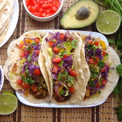 Three vegan potato tacos surrounded by diced tomatoes, avocado and lime halves, and homemade flour tortillas.