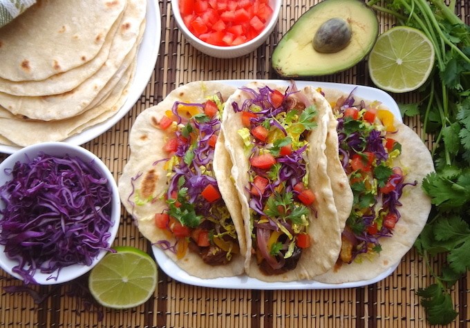 A top angle of 3 plated vegan potato tacos on a tan and brown mat. They're filled with seasoned refried beans, adobo seasoned potatoes, fajita style bell peppers and onions, fresh shredded purple cabbage and lettuce, cilantro leaves and diced tomatoes. Around the potato tacos sits a lime wedge, bowl of purple cabbage, plate of fresh homemade tortillas, a bowl of fresh diced tomatoes, half an avocado, and fresh cilantro.