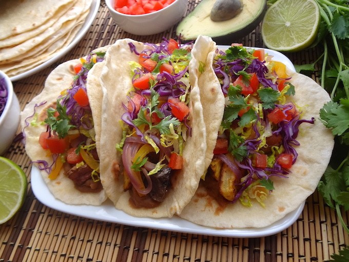 Siting on a tan and brown mat are 3 Vegan potato tacos on a plate. They're filled with seasoned refried beans, adobo seasoned potatoes, fajita style bell peppers and onions, fresh shredded purple cabbage and lettuce, and tomatoes. Around the potato tacos sits a lime wedge, bowl of purple cabbage, plate of fresh homemade tortillas, bowl of diced tomatoes, half an avocado, and fresh cilantro.