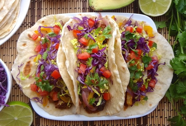 A close up top angle of 3 plated vegan potato tacos on a tan and brown mat. They're filled with seasoned refried beans, adobo seasoned potatoes, fajita style bell peppers and onions, fresh shredded purple cabbage and lettuce, and tomatoes. Around the potato tacos sits a lime wedge, bowl of purple cabbage, plate of fresh homemade tortillas, half an avocado, and fresh cilantro.