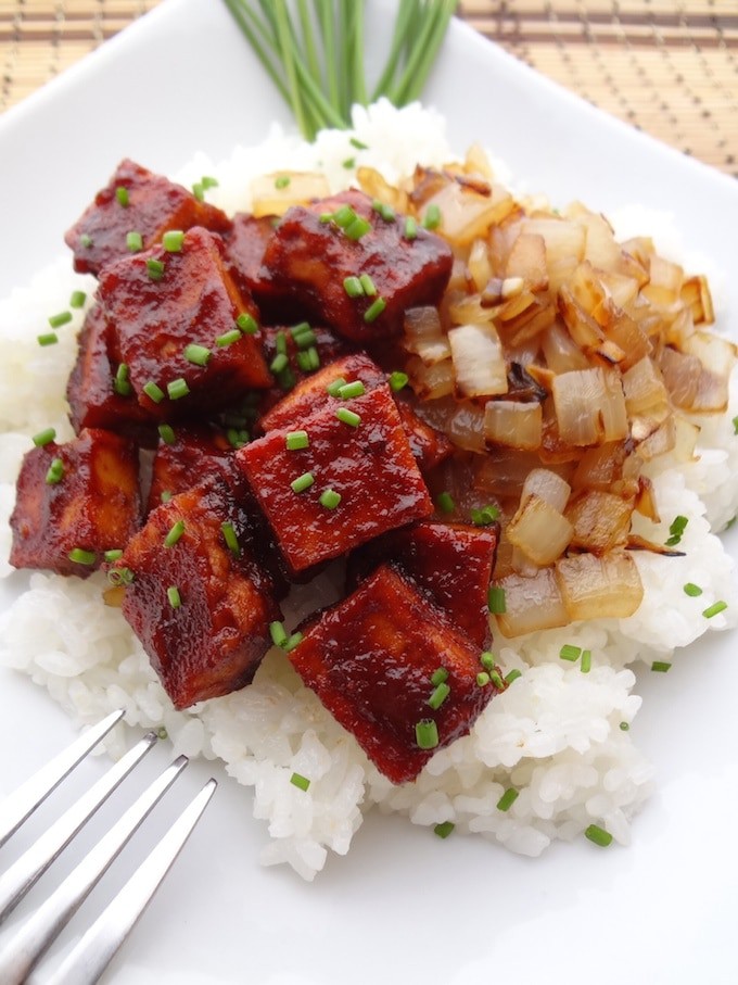 Baked BBQ tofu and caramelized onions on top of white sticky rice on a white plate. There is a fork to the bottom left. 