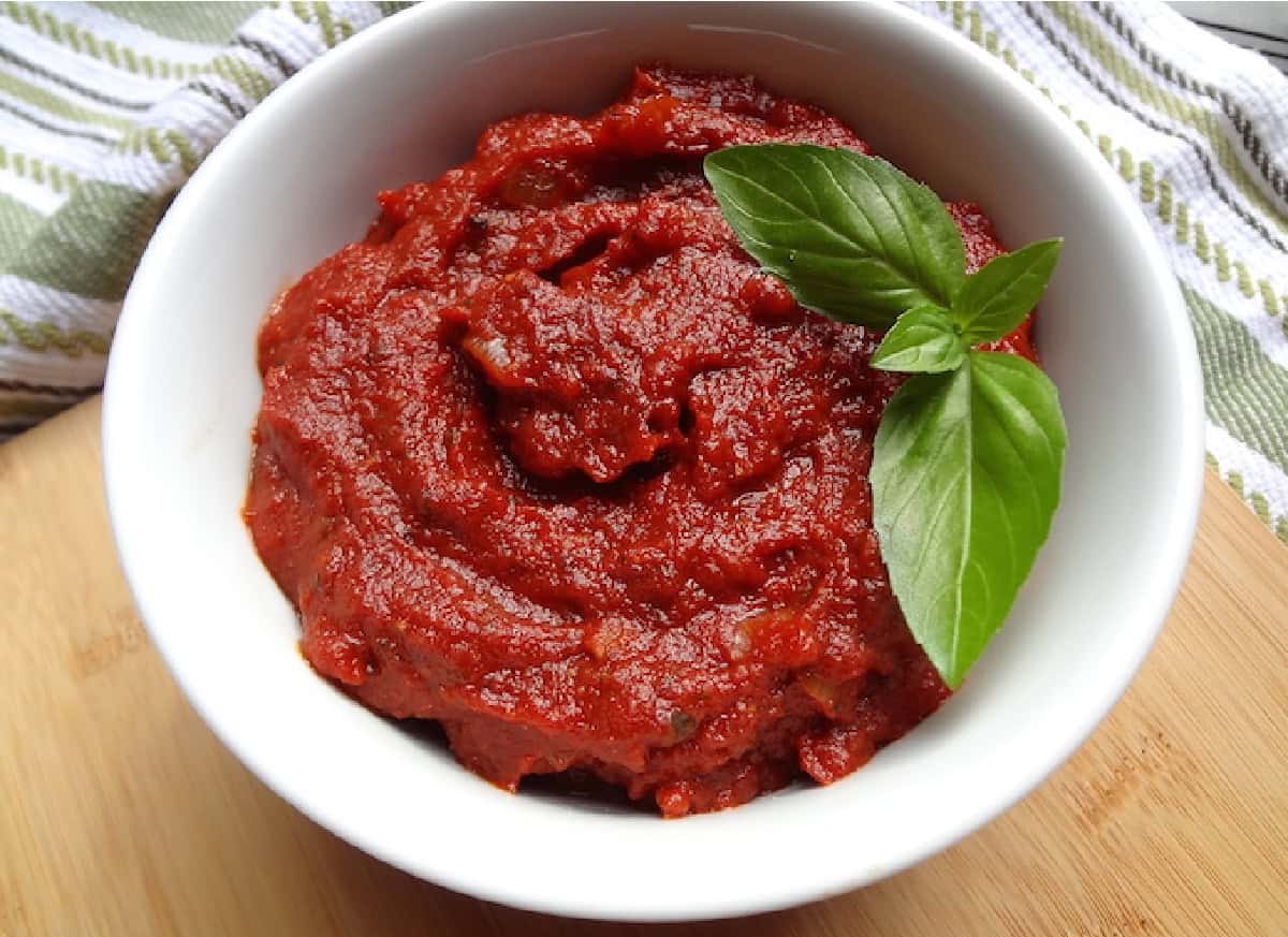A white bowl of marinara sauce with a fresh basil leaf.