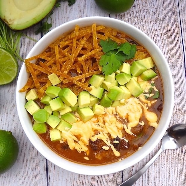 A bowl of vegan tortilla soup topped with cashew cheese, diced avocado, cilantro, and tortilla strips.