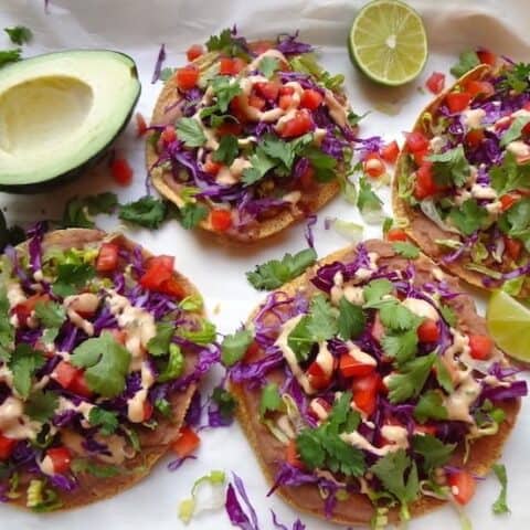Four vegan tostadas on a sheet of parchment paper alongside an avocado and lime cut in half.