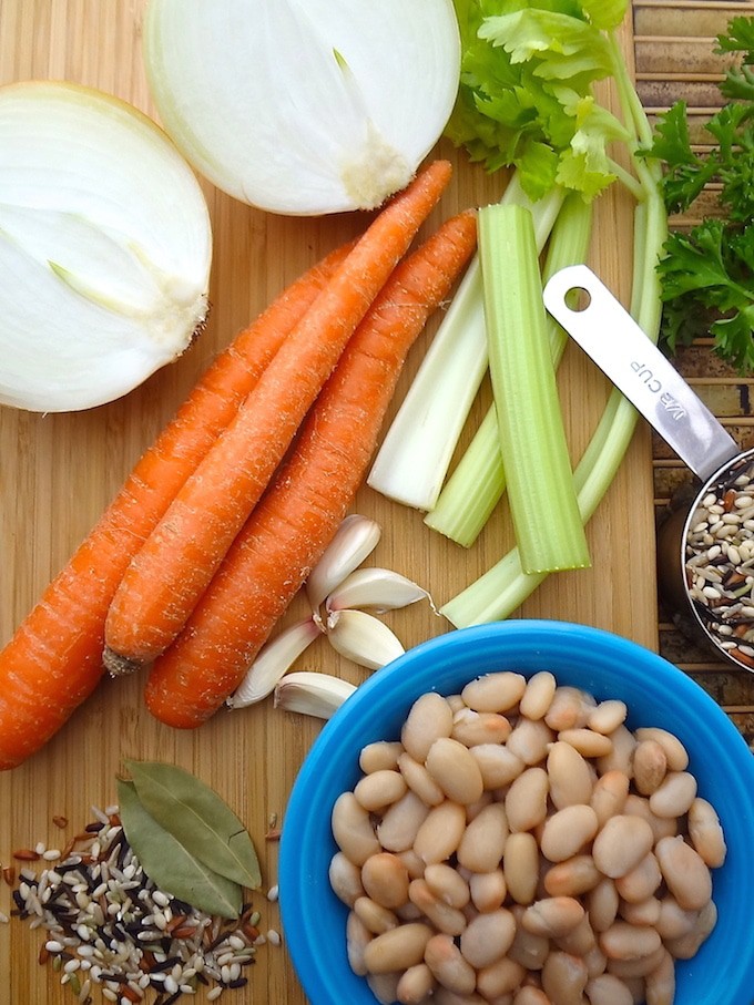 Quick One-Pot Veggie and Wild Rice Soup - vegan. Ingredients on a chopping board ready to be prepared for the soup. 3 carrots, an onion cut in half, celery stocks fresh parsley, garlic cloves, bay leaves, a blue bowl of white beans and a measuring cup of wild rice.