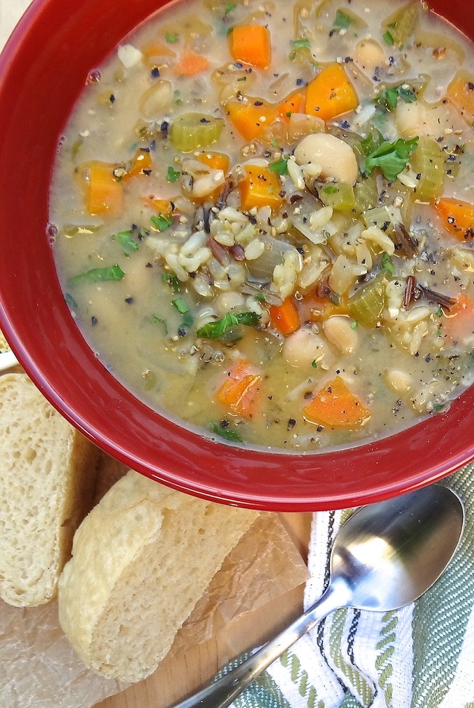 Quick One-Pot Veggie and Wild Rice Soup - vegan. A close up of a red bowl of veggie and wild rice soup. It's loaded with wild rice, white beans, chopped carrots & celery and diced onion then topped with fresh chopped parsley and black pepper. Served with a side of vegan fresh baked French bread slices for dipping.