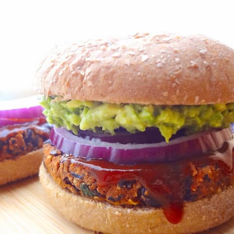 Vegan no crumble black bean burger on a wheat bun topped with mashed avocado, red onion slices and bbq sauce.