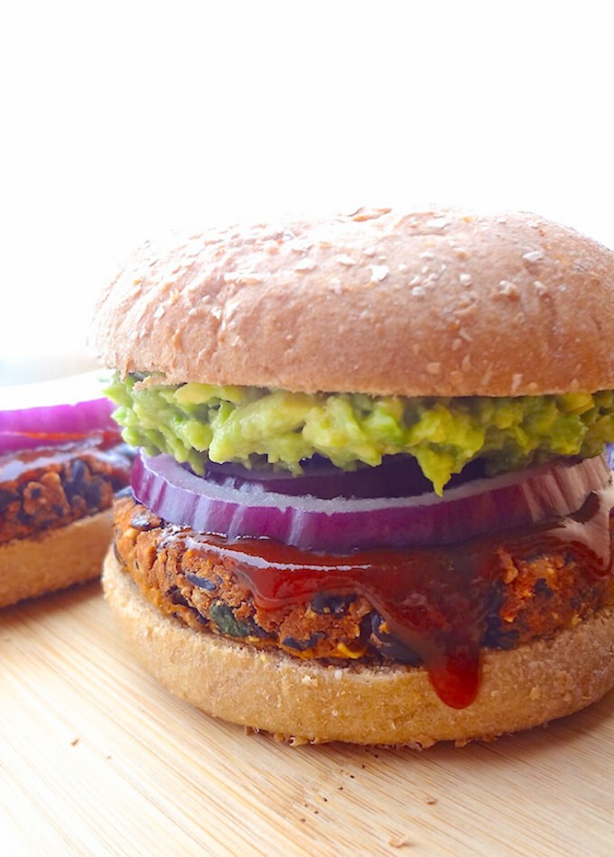 Vegan no crumble black bean burger on a wheat bun topped with mashed avocado, red onion slices and bbq sauce.