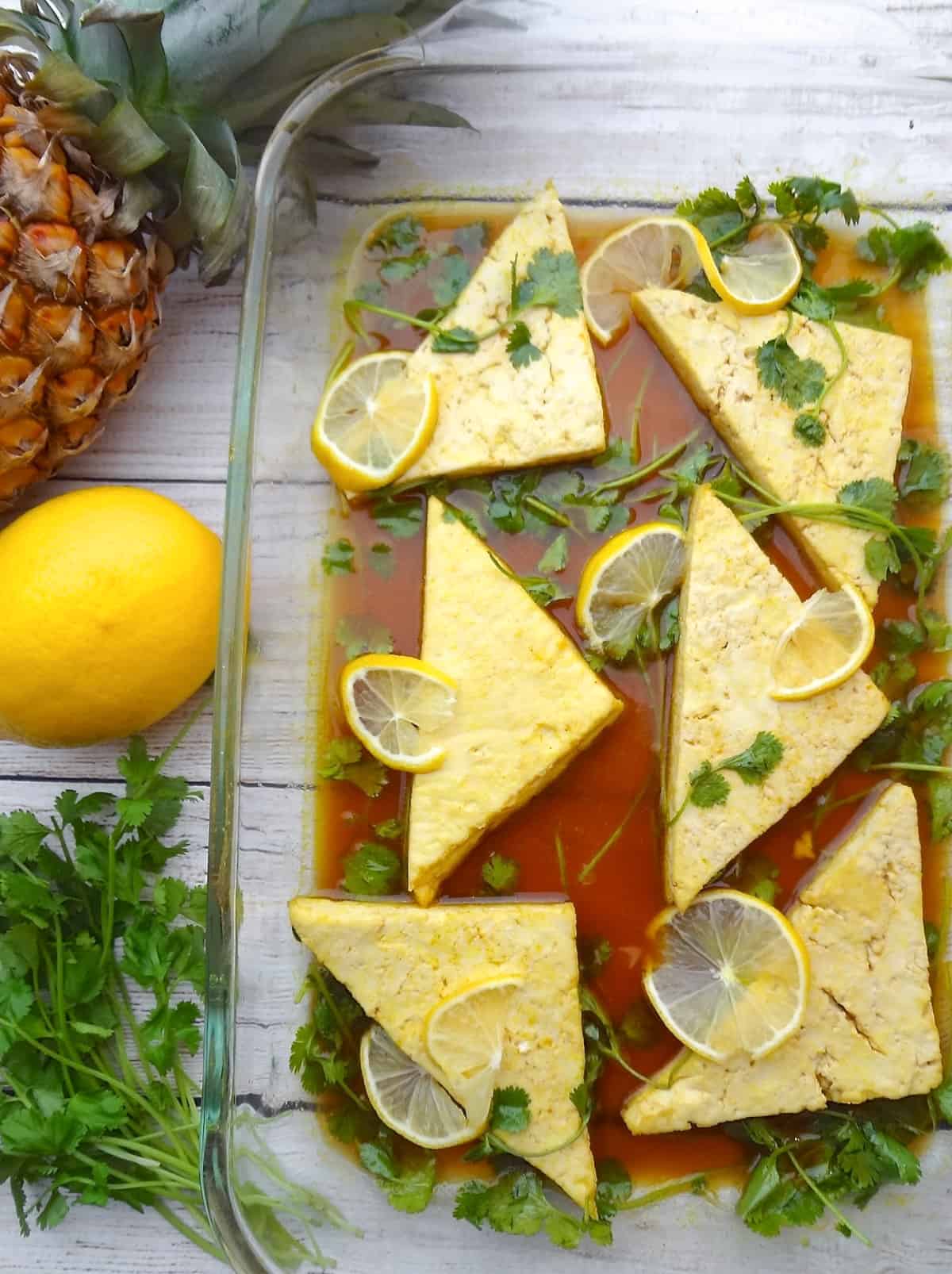 A glass baking dish with six triangle tofu filets in a lemon marinade with fresh cilantro and lemon slices on top.