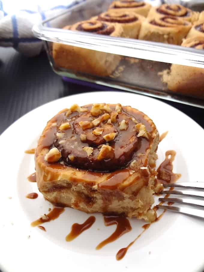 Refined sugar free vegan cinnamon roll on a white plate topped with maple icing and chopped walnuts.
