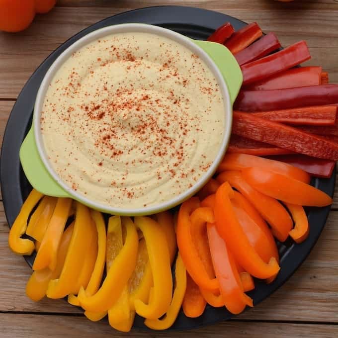 A bowl of hummus on a plate surrounded by yellow, orange, and red bell peppers.