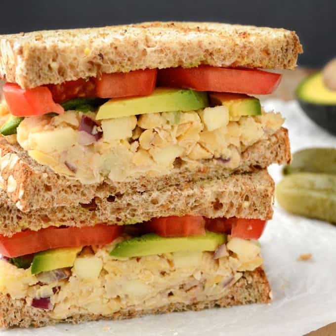 Apple walnut chickpea salad sandwich topped with avocado and tomato cut in half and stacked.
