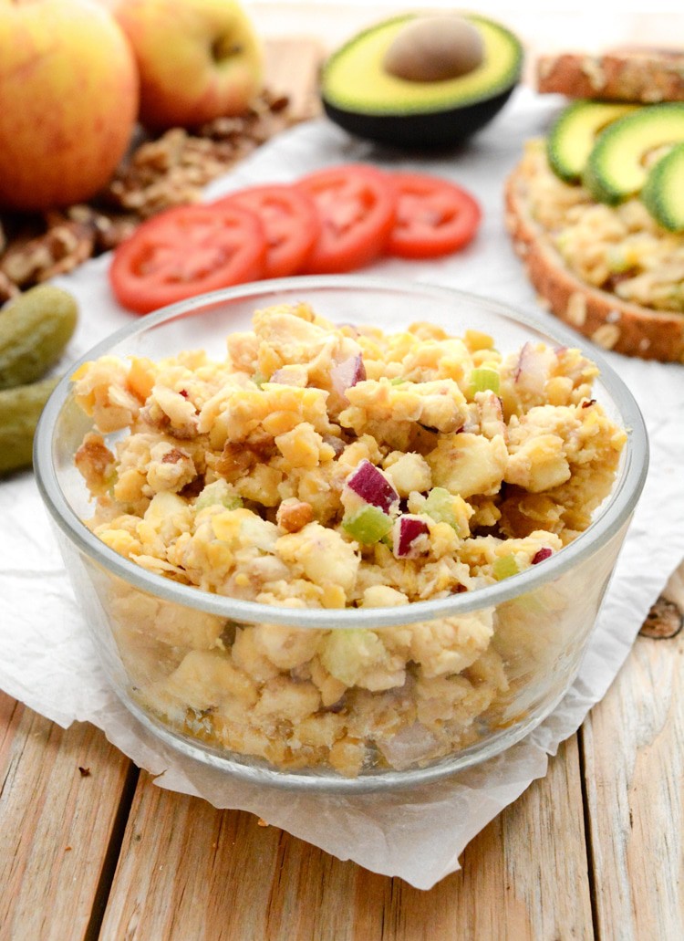 Apple-walnut chickpea salad in a bowl. It's on white parchment paper on raw wood. There are pickles, red and yellow gala apples, tomato slices, walnuts, a chickpea salad sandwich and half of an avocado in the back.