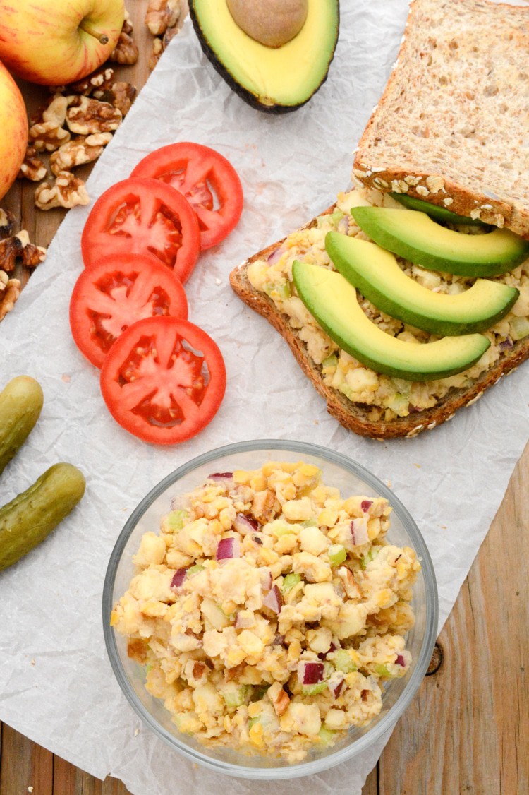 A top photo of the apple-walnut chickpea salad sandwich topped with avocado. The top bread is leaning against the back right corner of the sandwich. It's on white parchment paper on raw wood. There are also pickles, gala apples, tomato slices, walnuts, a bowl of chickpea salad and half of an avocado.