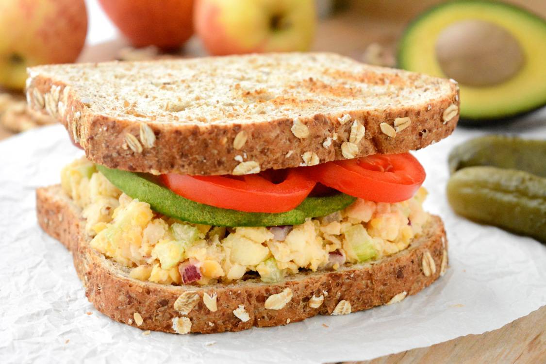 An apple-walnut chickpea salad sandwich topped with avocado and tomato slices. It's on white parchment paper. There are pickles, red and yellow gala apples, walnuts and half of an avocado in the back.