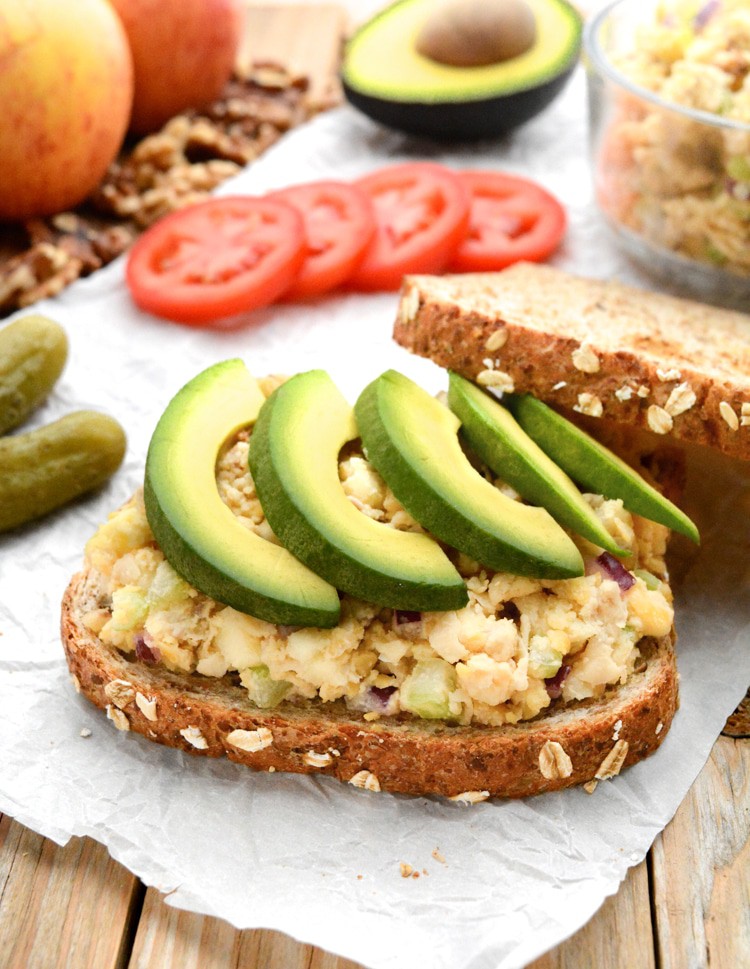 An apple-walnut chickpea salad sandwich topped with avocado. The top bread is leaning against the back right corner of the sandwich. It's on white parchment paper on raw wood. There are pickles, red and yellow gala apples, tomato slices, walnuts, a bowl of chickpea salad and half of an avocado in the back.