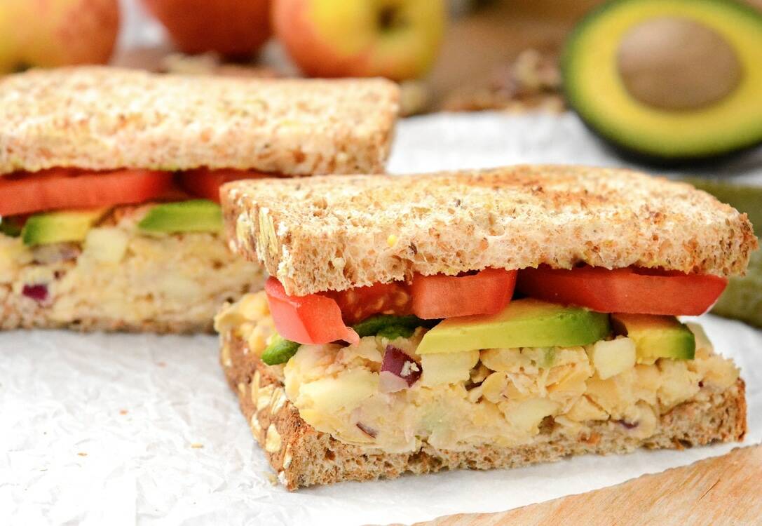 An apple-walnut chickpea salad sandwich topped with avocado and tomato slices. It's cut in half on white parchment paper. There are yellow and red apples, walnuts and half of an avocado in the back.