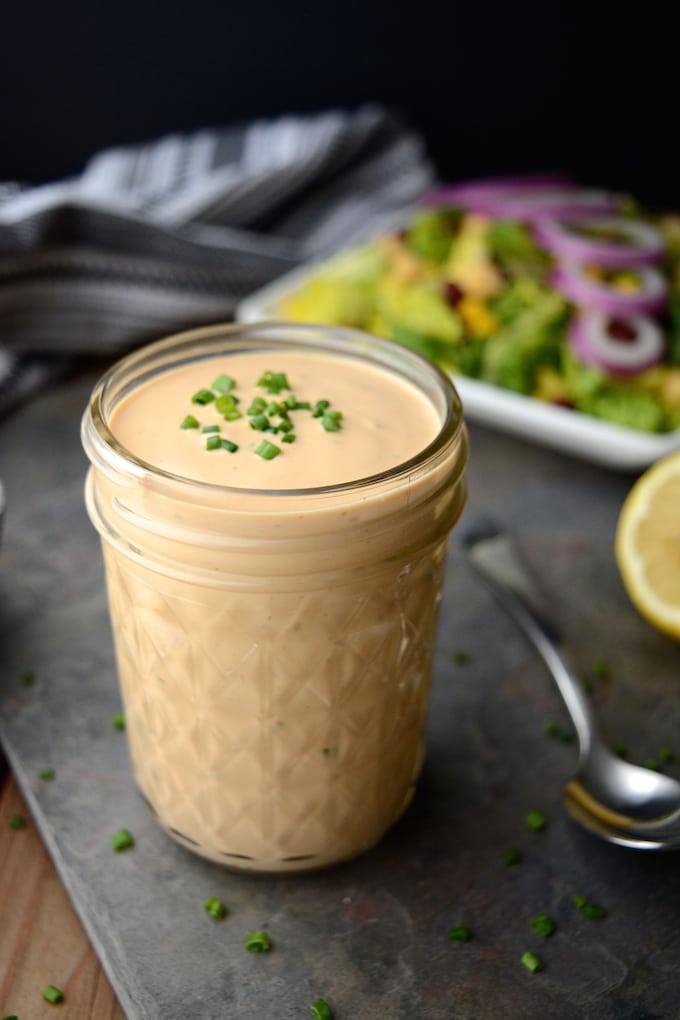 A jar of Creamy Vegan BBQ Ranch Dressing topped with green onions and a fresh salad in the background.