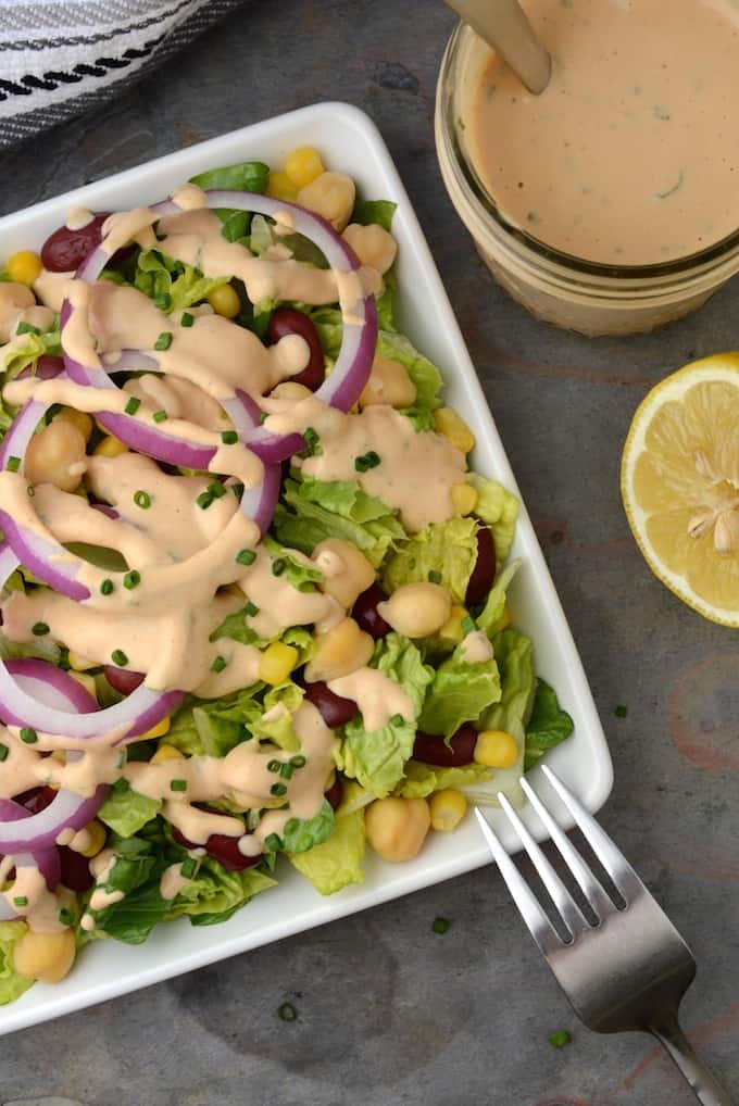 A salad topped with red onion rings, garbanzo beans, kidney beans, corn and creamy vegan BBQ Ranch Dressing with a jar of the dressing on the side.