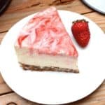 A slice of vegan strawberry swirl cheese cake and a strawberry on a white plate.