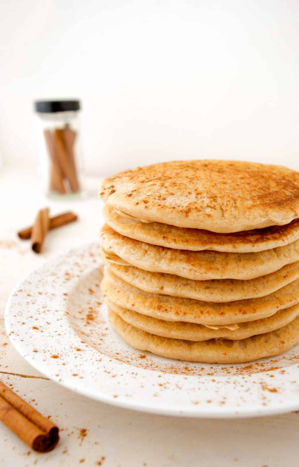 A stack of cinnamon pancakes on a white plate with a jar of cinnamon stick in the background.