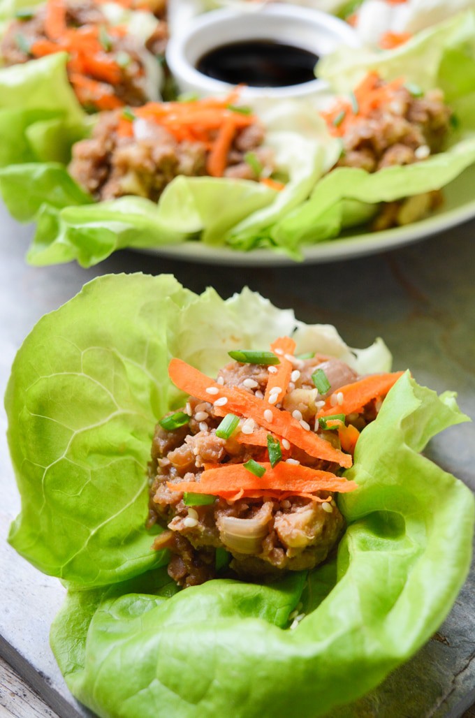 Vegan Lettuce Wraps-- Close up of the butter leaf lettuce filled with a lentil walnut mix, then topped with an Asian inspired sauce, grated carrots, green onions and toasted sesame seeds. More plated vegan lettuce wraps are in the back. They're served with an extra side of sauce in the middle of the plate.