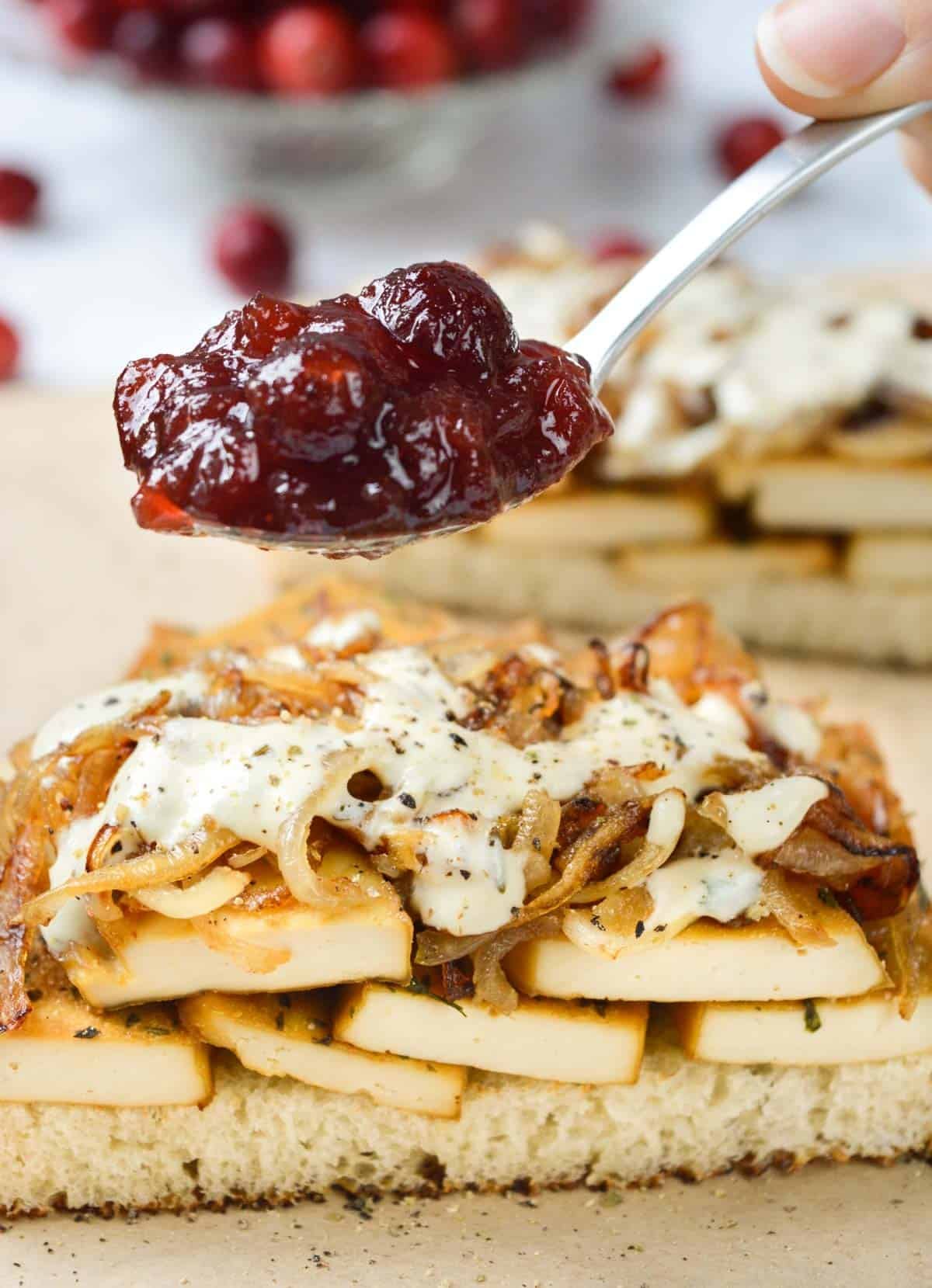 A scoop of cranberry sauce being added to a tofu, caramelized onion, and herb dressing sandwich.