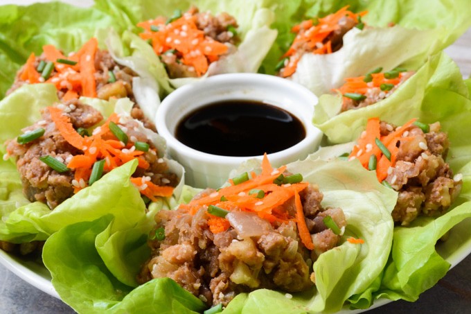 Vegan Lettuce Wraps-- Plated butter lettuce leaves filled with a lentil walnut mix, then topped with an Asian inspired sauce, grated carrots, green onions and toasted sesame seeds. Served with an extra side of sauce on the plate.