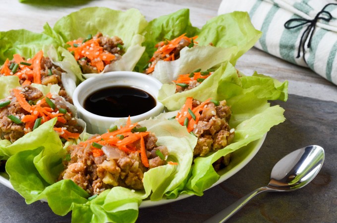Vegan Lettuce Wraps-- Plated butter lettuce leaves filled with a lentil walnut mix, then topped with an Asian inspired sauce, grated carrots, green onions and toasted sesame seeds. Served with an extra side of sauce on the plate and a spoon for drizzling.