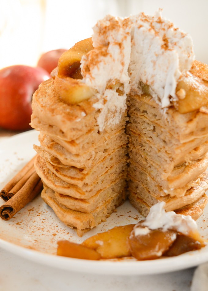 A stack of apple cinnamon pancakes topped with cinnamon apples and coconut whip. A triangle sized pieces has been cut out.
