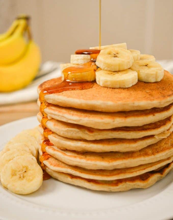 A stack of vegan banana milk pancakes with banana slices on top and syrup being poured on top.