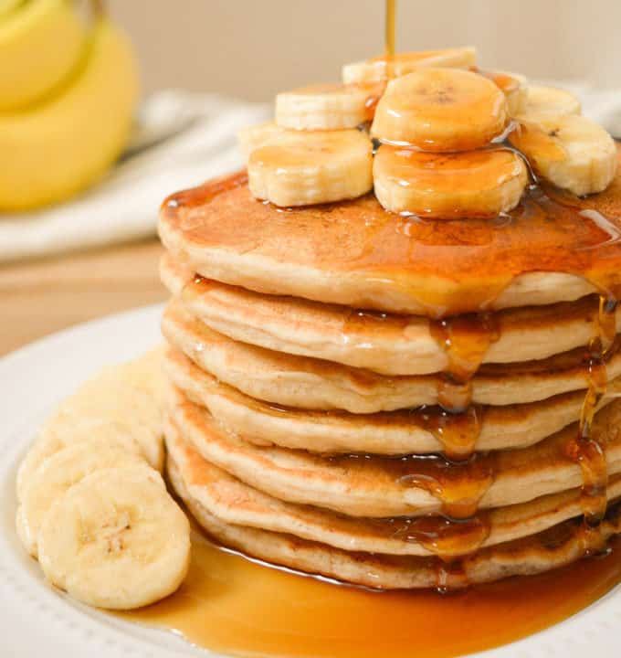 A stack of vegan banana milk pancakes with banana slices on top with maple syrup drizzled on top.