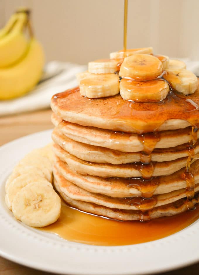 A stack of vegan banana milk pancakes with banana slices on top with maple syrup drizzled on top.