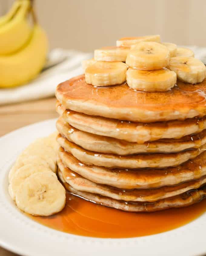 A stack of vegan banana milk pancakes with banana slices on top and maple syrup.