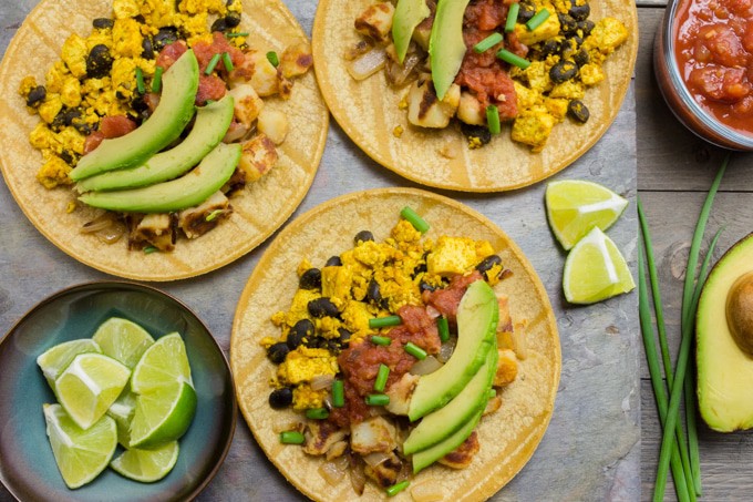 3 Vegan breakfast tacos on gray slate. There is a bowl of cut lime wedges, half an avocado and a bowl of salsa.