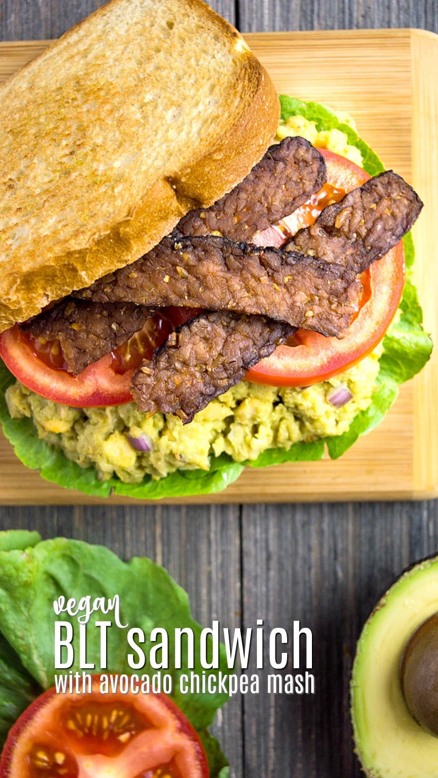 vegan BLT sandwich with avocado chickpea mash, tempeh bacon, and tomato slices on a cutting board.