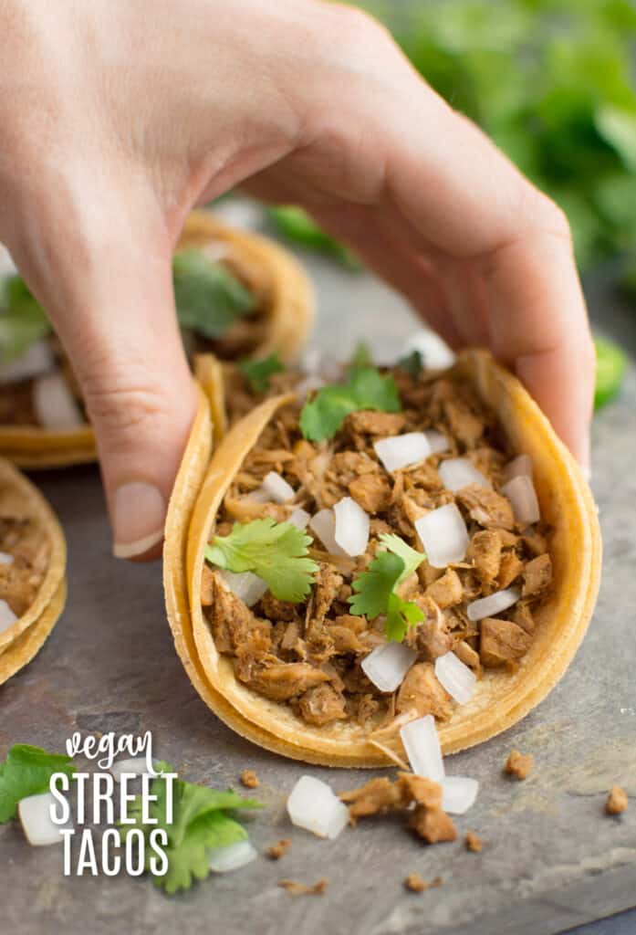 A hand grabbing a vegan taco on a corn tortilla filled with jackfruit, diced white onion and fresh cilantro.
