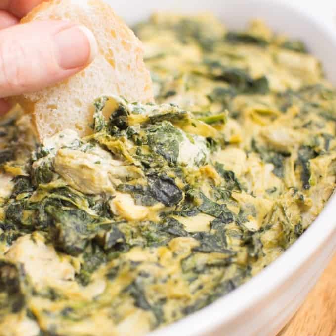 A hand dipping a piece of crusty bread into a dish of vegan spinach artichoke dip.