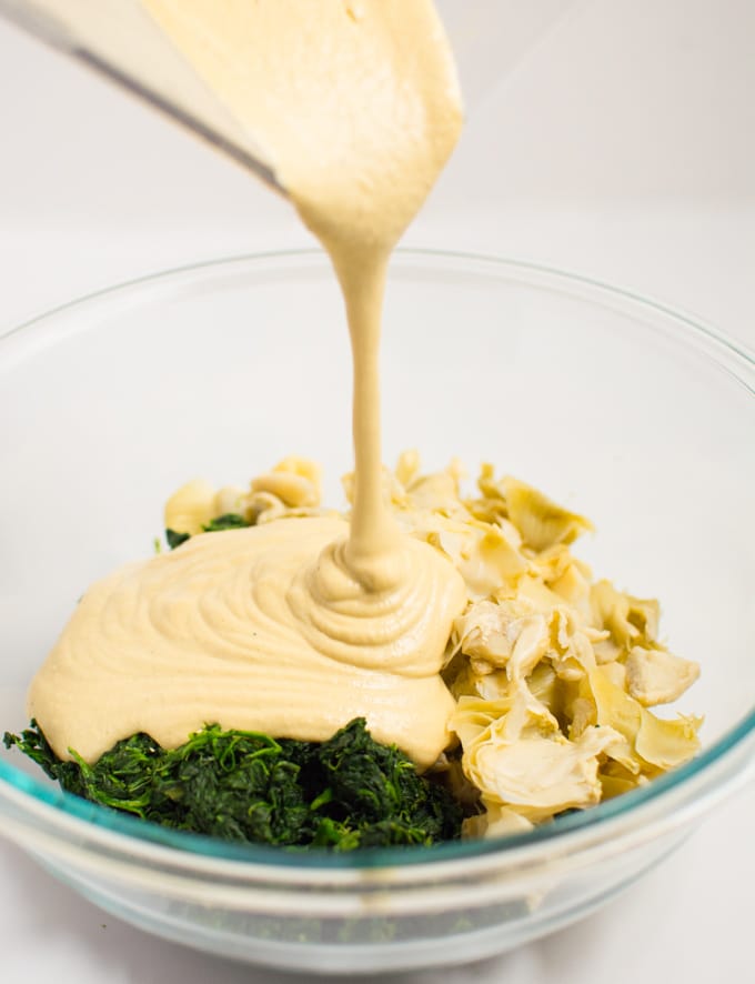 Cashew sauce being poured over spinach and artichoke hearts for vegan spinach artichoke dip.