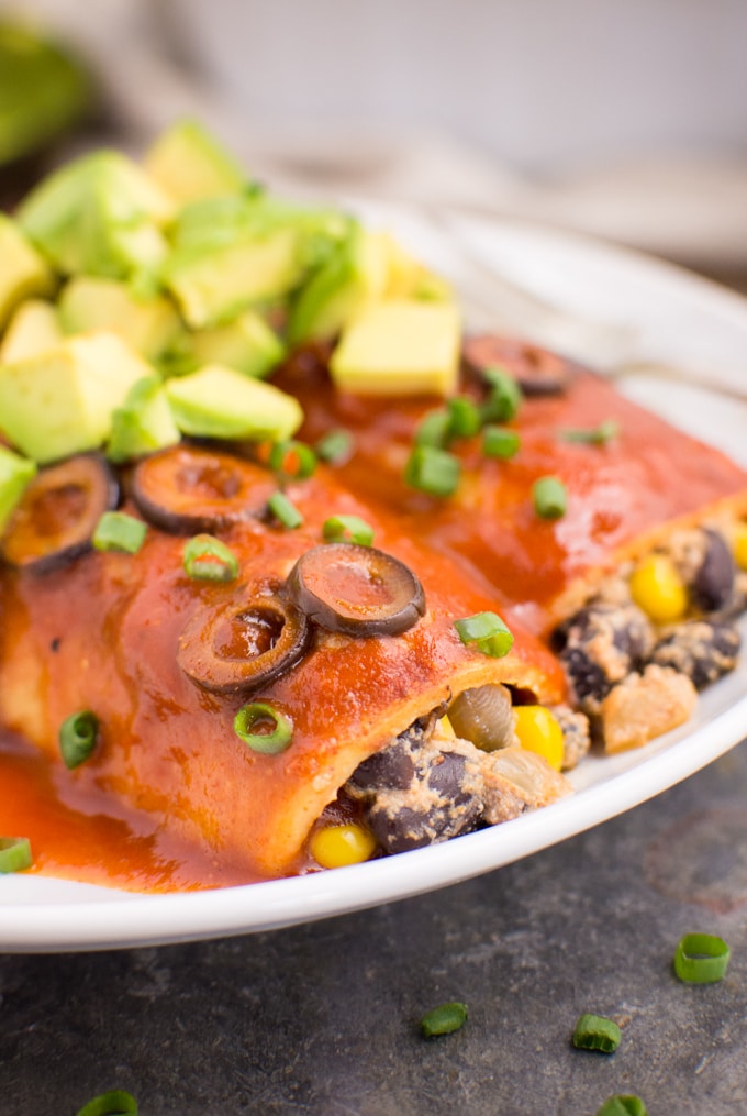 Two vegan black bean enchiladas on a white plate topped with extra sauce, green onions, sliced olives, and diced avocado.