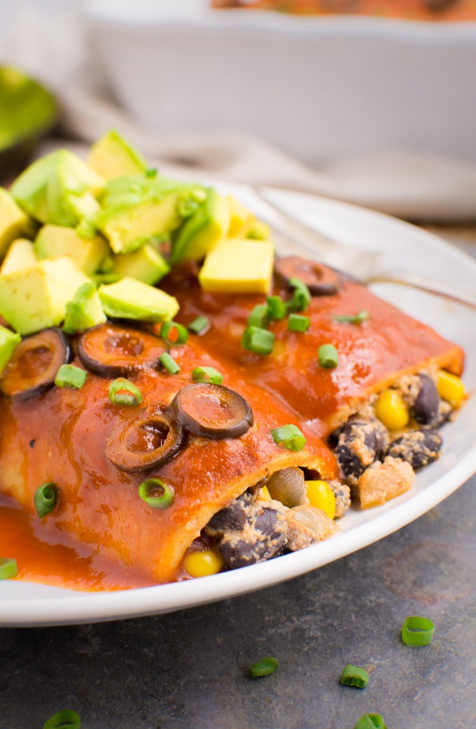 2 vegan enchiladas on a white plate stuffed with a creamy black bean filling then topped with sliced olives, green onions and diced avocado.