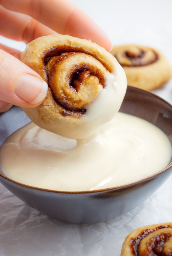 A mini cinnamon roll being dipped in a small bowl of dairy-free icing.