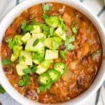 Vegan chili in a white bowl topped with diced avocado, chopped cilantro and jalapeño slices.
