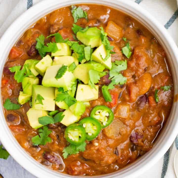 Vegan chili in a white bowl topped with diced avocado, chopped cilantro and jalapeño slices.