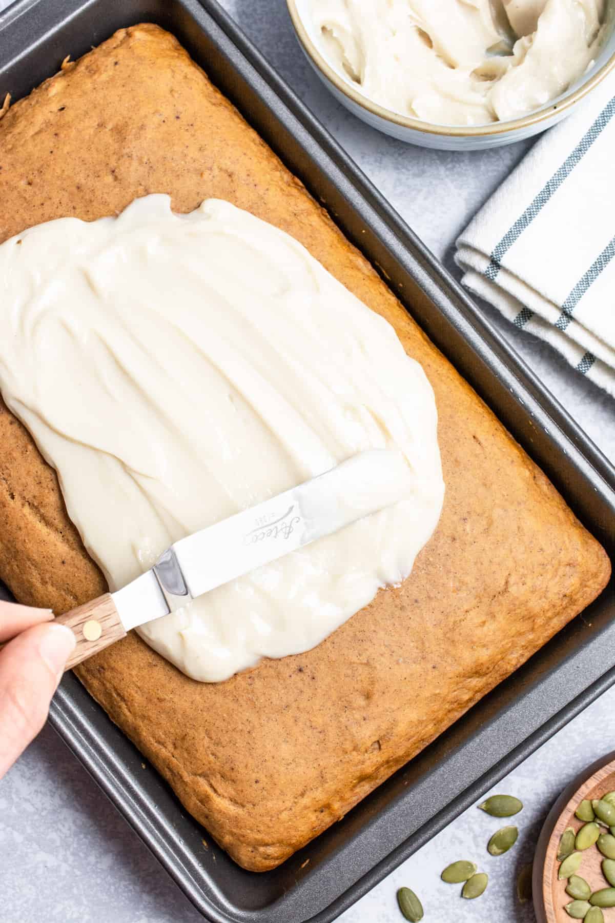 A hand using a spatula to spread frosting on a rectangle shaped cake.