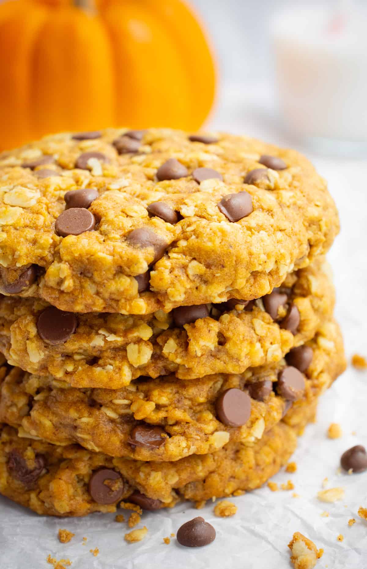 A stack of vegan pumpkin oatmeal cookies with chocolate chips, a glass of almond milk, and an orange pumpkin in the background.