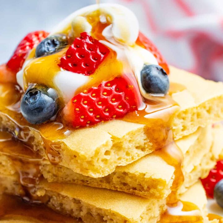 A stack of sheet pan pancakes topped with fruit, whip, and maple syrup.