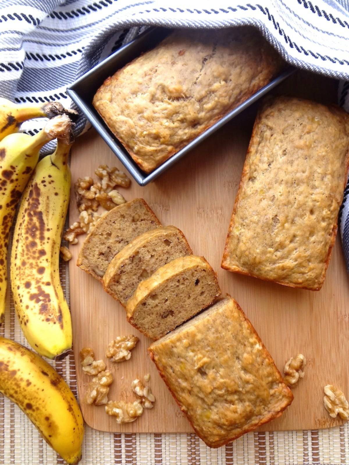 3 loaves of vegan banana bread on a cutting board.  One in the loaf pan, one whole, and the other with 3 slices cut.