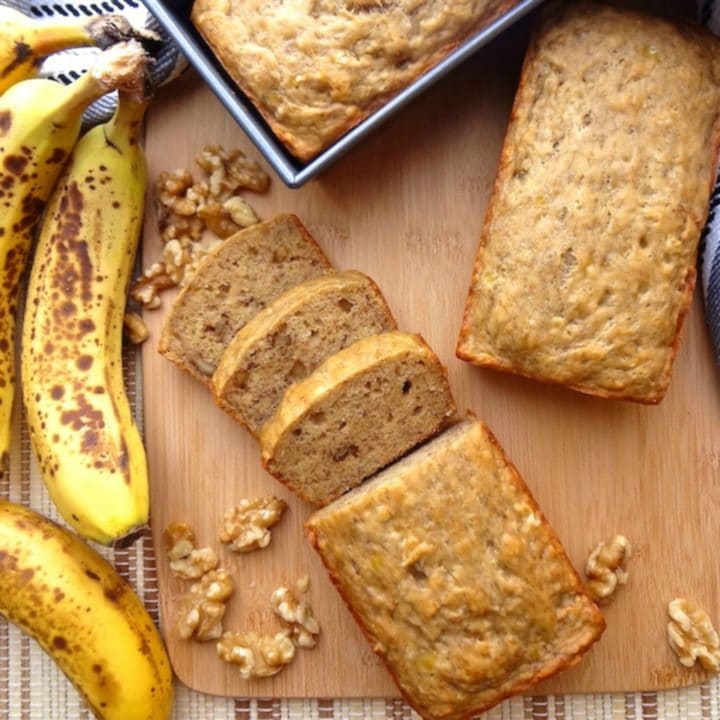 A mini loaf of vegan banana bread alongside ripe bananas and chopped walnuts.